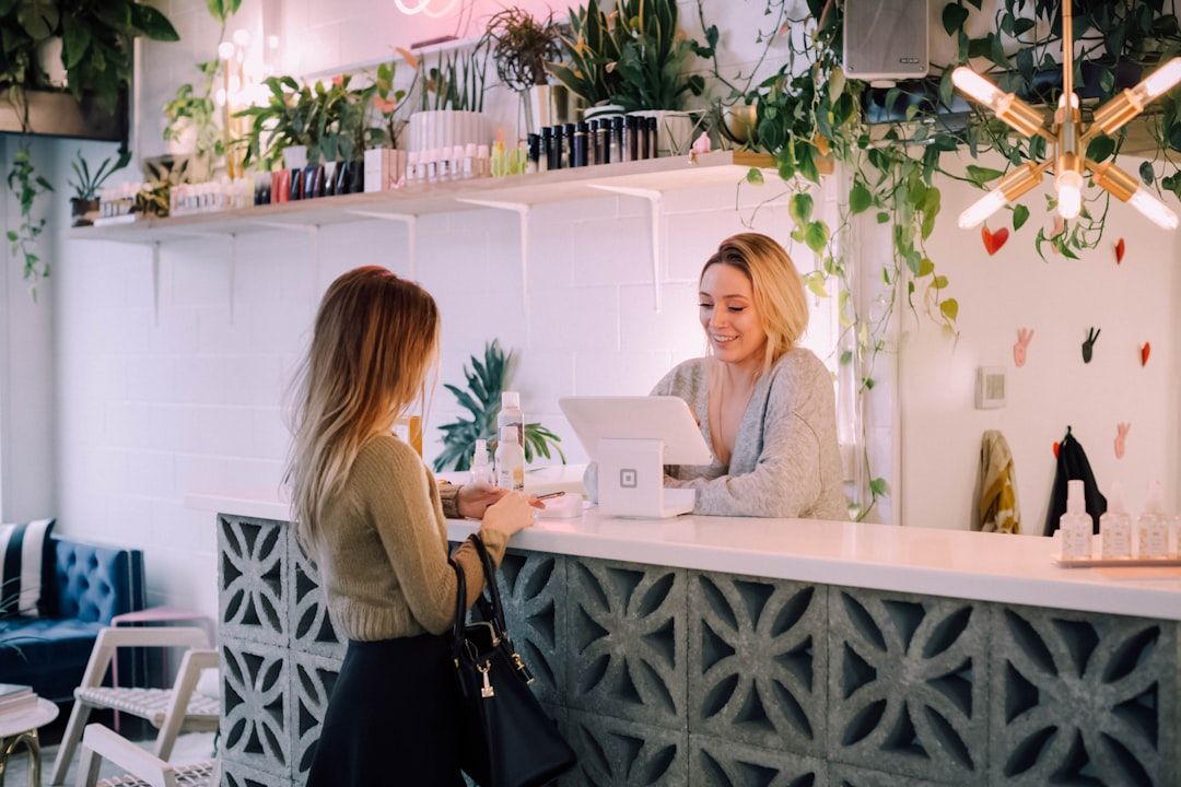 Recepcionista sonriente atendiendo a una clienta en el mostrador de un salón con POS y plantas; Descubre dónde trabaja un recepcionista y cómo iniciar tu carrera en este emocionante campo laboral.