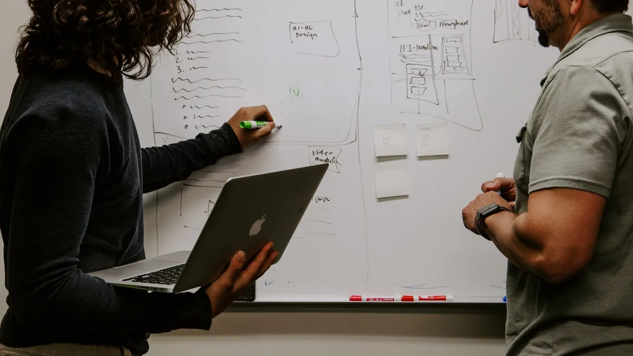 Un grupo de personas sentadas en círculo, con un coach liderando la conversación y apuntando a un pizarrón con metas escritas 